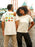 Two people wearing white t-shirts with colorful graphics in front of a carnival.