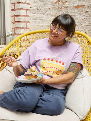 A person sitting in a yellow woven chair wearing a light purple graphic t-shirt with a birthday cake design, blue sweatpants, and holding a plate with a slice of cake.