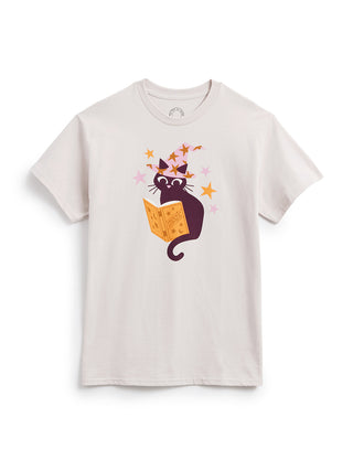 A light beige crewneck t-shirt featuring a cartoon graphic of a black cat reading an orange magic book, surrounded by pink and orange stars, displayed on a clean white background.