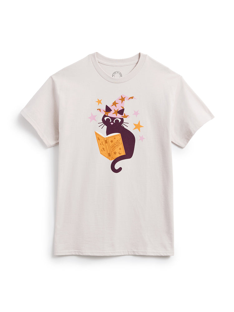 A light beige crewneck t-shirt featuring a cartoon graphic of a black cat reading an orange magic book, surrounded by pink and orange stars, displayed on a clean white background.