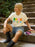 A woman sits on outdoor stone steps wearing a cream graphic T-shirt that reads 'BOOK SCHLEPPER' in multicolored letters, paired with denim shorts and black boots, with a stack of books beside her.