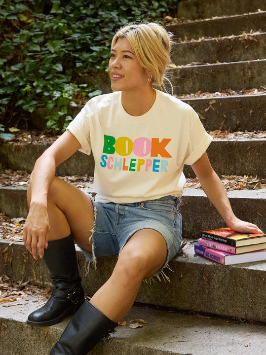 A woman sits on outdoor stone steps wearing a cream graphic T-shirt that reads 'BOOK SCHLEPPER' in multicolored letters, paired with denim shorts and black boots, with a stack of books beside her.