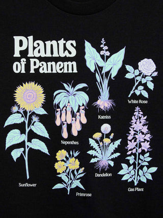 A close-up of a black t-shirt featuring a graphic print of various plants labeled 'Plants of Panem' including a sunflower, katriss, white rose, nepenthes, dandelion, primrose, and gas plant.