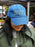 A person wearing a blue denim baseball cap with yellow embroidered text that reads 'YOU'RE GOING TO MESS UP AND IT'S GOING TO BE OKAY' in an outdoor market setting.