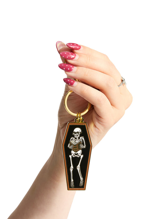 Hand holding a keychain with a skeleton design in a coffin on a white background