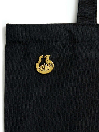 A close-up of a black canvas tote bag featuring a small gold sunrise on the reaping enamel pin on the front, set against a plain white background.