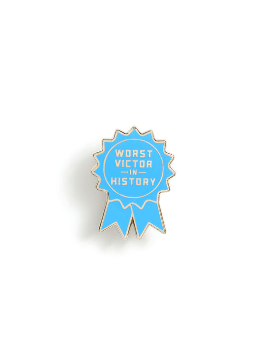 A light blue ribbon-shaped enamel pin with gold metal edging and the text 'WORST VICTOR IN HISTORY' in white, shown on a plain white background.