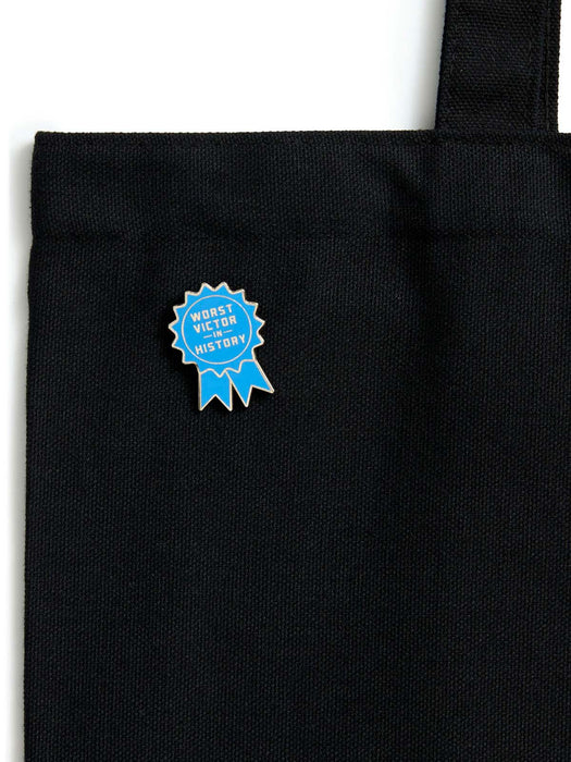 A close-up of a black canvas tote bag with a blue and gold enamel pin on the front that reads 'WORST VICTOR IN HISTORY'.
