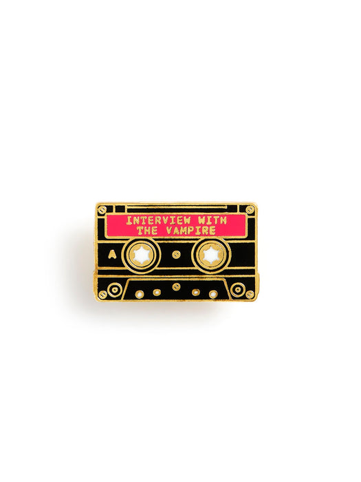 A cassette tape-shaped enamel pin with a black body, gold metal outlines, a bright pink title bar reading 'INTERVIEW WITH THE VAMPIRE', and two circular reel holes with white star details.