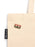 A close-up of a beige canvas tote bag with a small cassette tape enamel pin on the side. The pin has pink, gold, and black details.