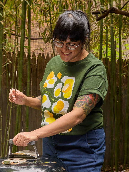 A woman wearing a green graphic t-shirt with a yellow and white fried egg print, blue jeans, and glasses is smiling while holding the lid of a grill in front of a bamboo fence.
