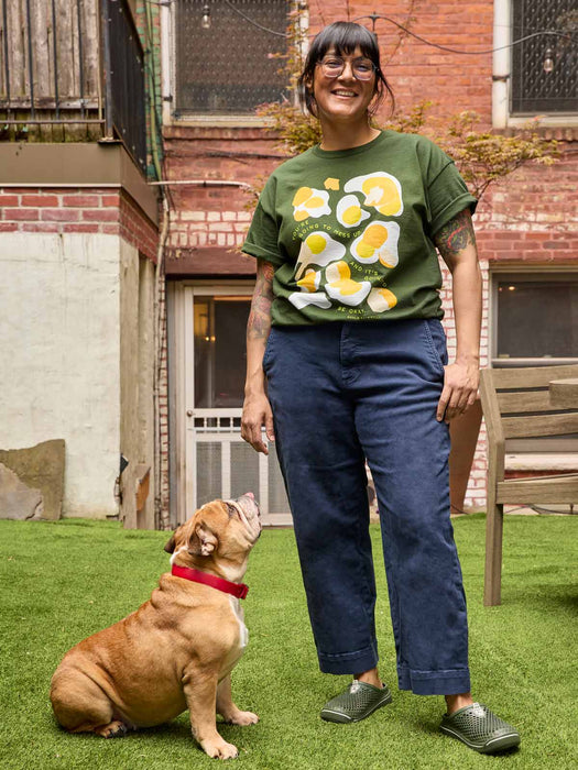A woman stands on green grass in front of a brick building, wearing a dark green graphic t-shirt with a yellow and white egg pattern, navy blue pants, and green clogs. A brown dog with a red collar sits beside her looking up.