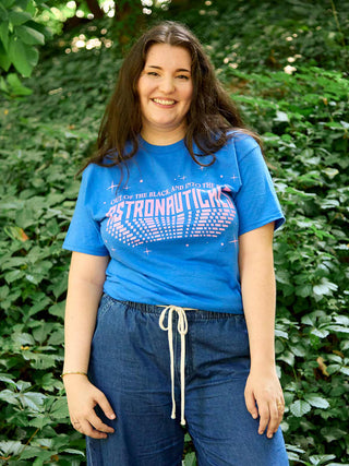 A smiling woman with long dark hair stands outdoors in front of a green leafy background, wearing a bright blue graphic T-shirt that reads 'OUT OF THE BLACK AND INTO THE BLUE ASTRONAUTICALS' in pink and white, paired with dark blue drawstring pants.