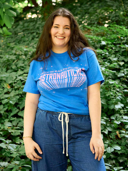 A smiling woman with long dark hair stands outdoors in front of a green leafy background, wearing a bright blue graphic T-shirt that reads 'OUT OF THE BLACK AND INTO THE BLUE ASTRONAUTICALS' in pink and white, paired with dark blue drawstring pants.