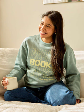 Woman wearing a light blue sweatshirt with 'BOOKS' printed on it, sitting on a couch holding a mug.