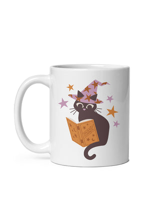 A white ceramic mug featuring a cartoon illustration of a black cat wearing a purple witch hat and reading an orange magic book, surrounded by small stars.