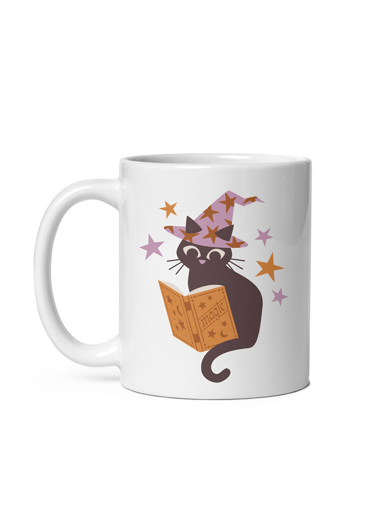 A white ceramic mug featuring a cartoon illustration of a black cat wearing a purple witch hat and reading an orange magic book, surrounded by small stars.