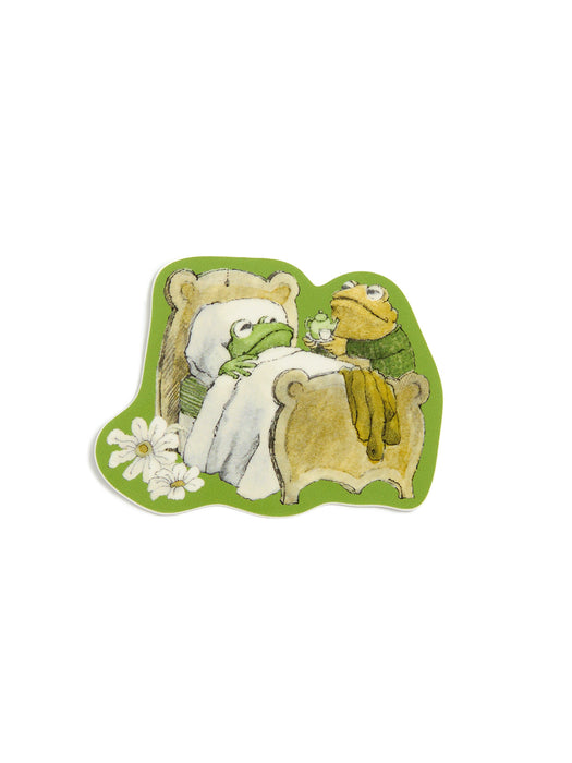 A sticker featuring a whimsical illustration of a green frog in bed with a white blanket set against a light green background with white daisy flowers in the lower left corner and Toad holding a tea kettle.