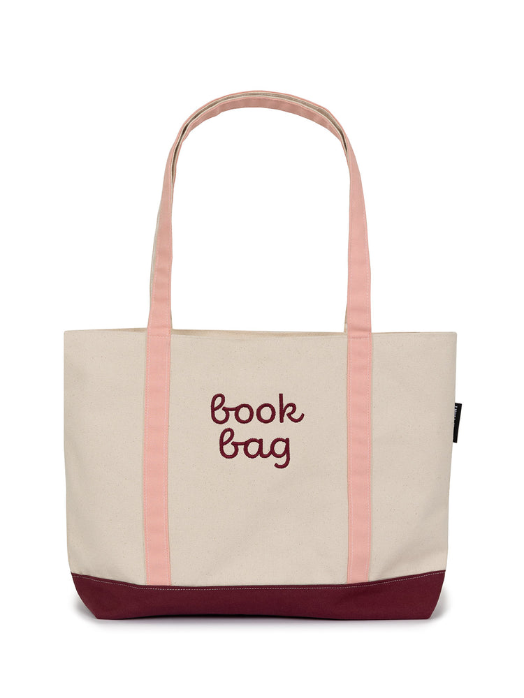 A large zippered tote bag made of canvas with peaches and cream colored straps and burgundy bottom, labeled 'book bag'.