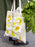Beige tote bag with yellow floral design held by a person with greenery in the background