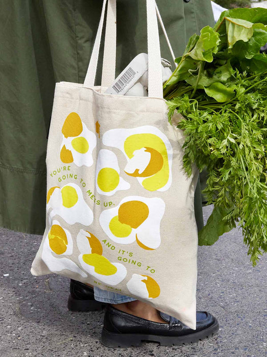Beige tote bag with yellow floral design held by a person with greenery in the background