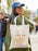 Person holding a tote bag with a cake design and text, walking outdoors.