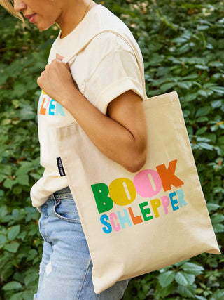 A person outdoors carrying a cream canvas tote bag with the text 'BOOK SCHLEPPER' printed in colorful letters. The person is wearing a t-shirt and light blue jeans, with green foliage in the background.