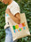 A person outdoors carrying a cream canvas tote bag with the text 'BOOK SCHLEPPER' printed in colorful letters. The person is wearing a t-shirt and light blue jeans, with green foliage in the background.