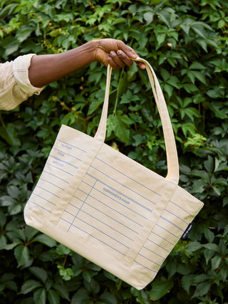 A person's hand holding a cream-colored canvas tote bag with blue lined writing sections, set against a background of green leafy vines.