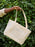 A person's hand holding a cream-colored canvas tote bag with blue lined writing sections, set against a background of green leafy vines.