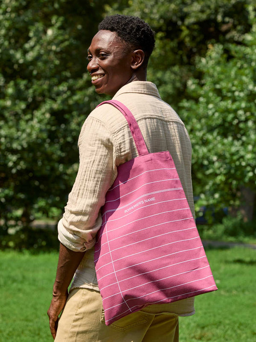 A smiling man in a beige shirt and khaki pants is shown from the back in an outdoor park setting, carrying a maroon red canvas library tote bag with thin white horizontal stripes and white text that reads 'BORROWER'S NAME'.