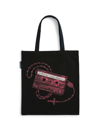A black canvas tote bag with black shoulder straps, featuring a graphic print of a cassette tape with a rosary chain and cross in a dusty pink color, set against a plain white background.