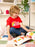 A young child with curly blonde hair sits cross-legged on a patchwork blanket in a bright room, smiling while reading a colorful children's book. The child is wearing a red graphic T-shirt that reads 'Reading Rainbow' with a rainbow design, blue jeans, and gray socks.