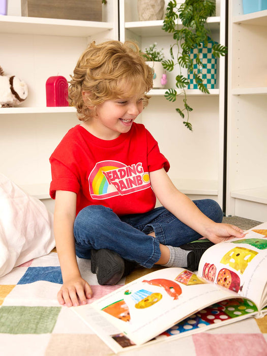 A young child with curly blonde hair sits cross-legged on a patchwork blanket in a bright room, smiling while reading a colorful children's book. The child is wearing a red graphic T-shirt that reads 'Reading Rainbow' with a rainbow design, blue jeans, and gray socks.