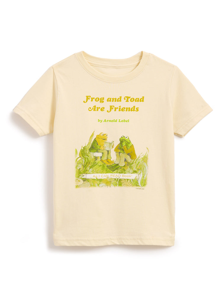 A cream-colored short-sleeve t-shirt featuring a graphic print of the 'Frog and Toad Are Friends' book cover, including the title in yellow text and the illustration below, displayed on a clean white background.